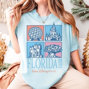 May include: Light blue t-shirt featuring four square images of Florida landmarks. The shirt includes the text "FLORIDA!!!" and "Walt Disney World" in a script font. The images include a sphere, a castle, a building, and a tree.
