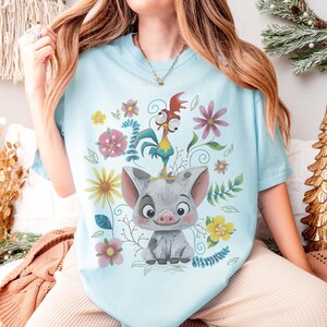 Puede incluir: Camiseta azul claro con un dise&ntilde;o de cerdo y gallo de dibujos animados, rodeada de flores y hojas coloridas. La ilustraci&oacute;n es juguetona y brillante, ideal para los fan&aacute;ticos de las pel&iacute;culas de animaci&oacute;n.