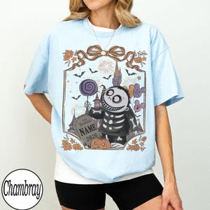 K&ouml;nnte beinhalten: Hellblaues T-Shirt mit einem Halloween-Motiv. Das Design zeigt eine Skelettfigur, die einen Lolli h&auml;lt, einen Grabstein mit dem Text "NAME 2026" und andere Halloween-Elemente. Das T-Shirt hat kurze &Auml;rmel und einen Rundhalsausschnitt.