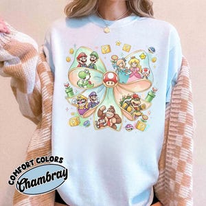Op de afbeelding: Lichtblauw Comfort Colors Chambray T-shirt met een kleurrijke afbeelding van Super Mario-personages in een strikvorm. Het ontwerp bevat Mario, Luigi, Yoshi, Princess Peach en Bowser, met vraagtekendozen en munten.