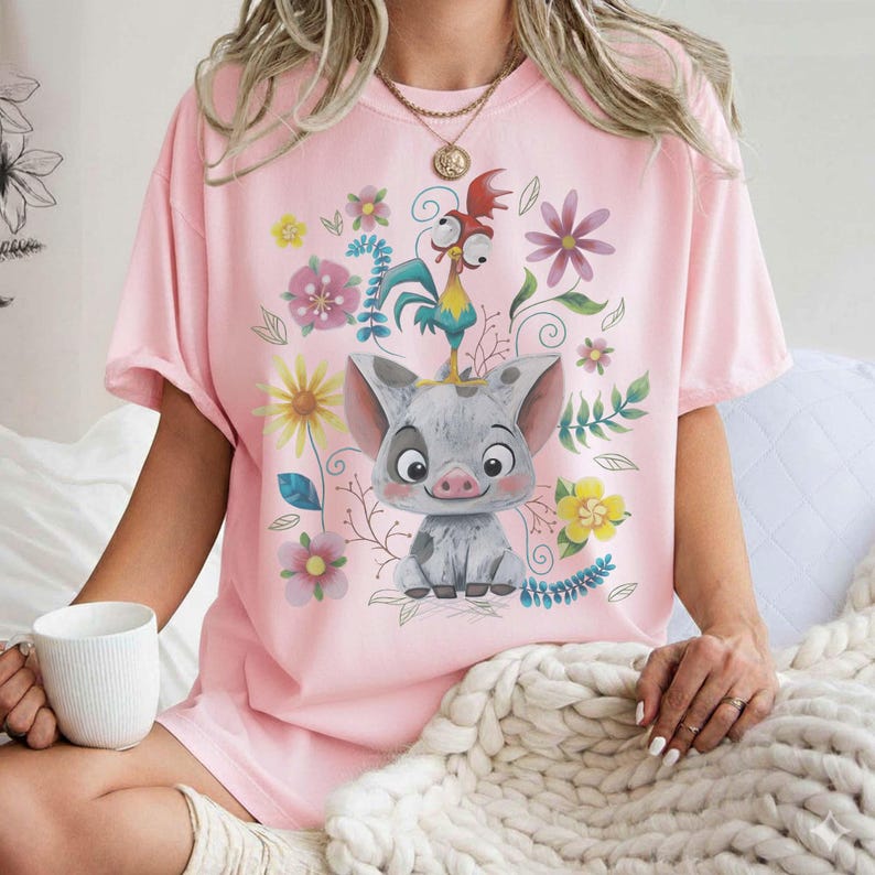 Puede incluir: Camiseta rosa claro con un dise&ntilde;o de cerdo de dibujos animados y un gallo, rodeado de flores y follaje coloridos. El dise&ntilde;o est&aacute; centrado en la parte delantera. La camiseta tiene mangas cortas y un corte holgado.