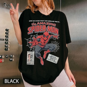 K&ouml;nnte beinhalten: Schwarzes T-Shirt mit einer Comic-Grafik von Spider-Man. Das Design enth&auml;lt den Text "the AMAZING SPIDER-MAN" und "NEVER HAS ANYONE FOUGHT SUCH MERCILESS FOES AS..." sowie ein Bild von Peter Parker.