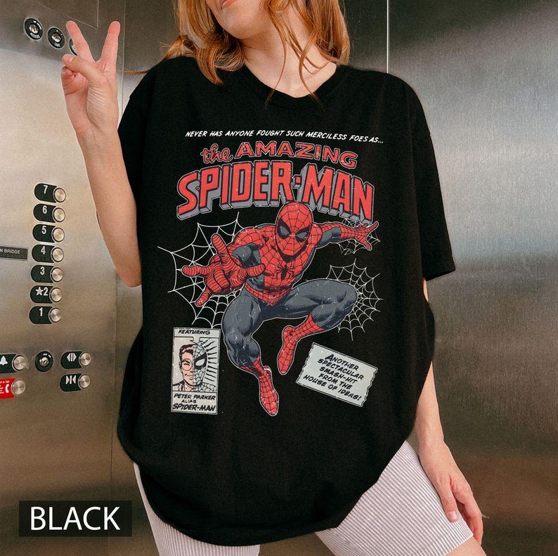 K&ouml;nnte beinhalten: Schwarzes T-Shirt mit einer Comic-Grafik von Spider-Man. Das Design enth&auml;lt den Text "the AMAZING SPIDER-MAN" und "NEVER HAS ANYONE FOUGHT SUCH MERCILESS FOES AS..." sowie ein Bild von Peter Parker.