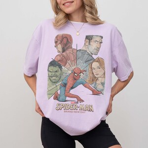 May include: A lilac t-shirt with a graphic of Spider-Man and other Marvel characters. The design includes the text "SPIDER-MAN BRAND NEW DAY". The shirt is a crew neck and has short sleeves.