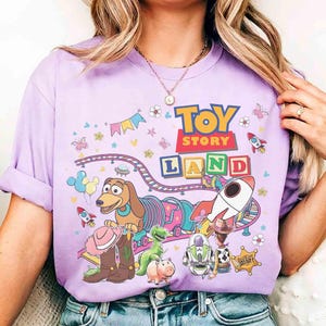 May include: A lilac t-shirt with a vibrant "Toy Story Land" design. The graphic showcases characters such as Slinky Dog, Buzz Lightyear, and Woody, alongside rockets, flowers, and a train track. The words "TOY STORY LAND" are clearly visible.
