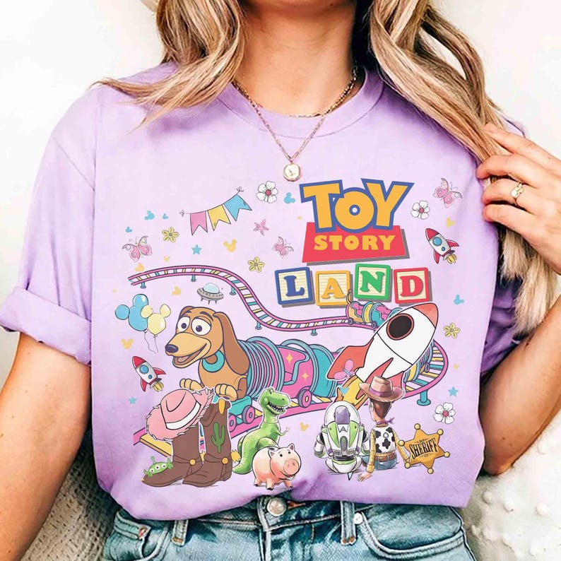 May include: A lilac t-shirt with a vibrant "Toy Story Land" design. The graphic showcases characters such as Slinky Dog, Buzz Lightyear, and Woody, alongside rockets, flowers, and a train track. The words "TOY STORY LAND" are clearly visible.