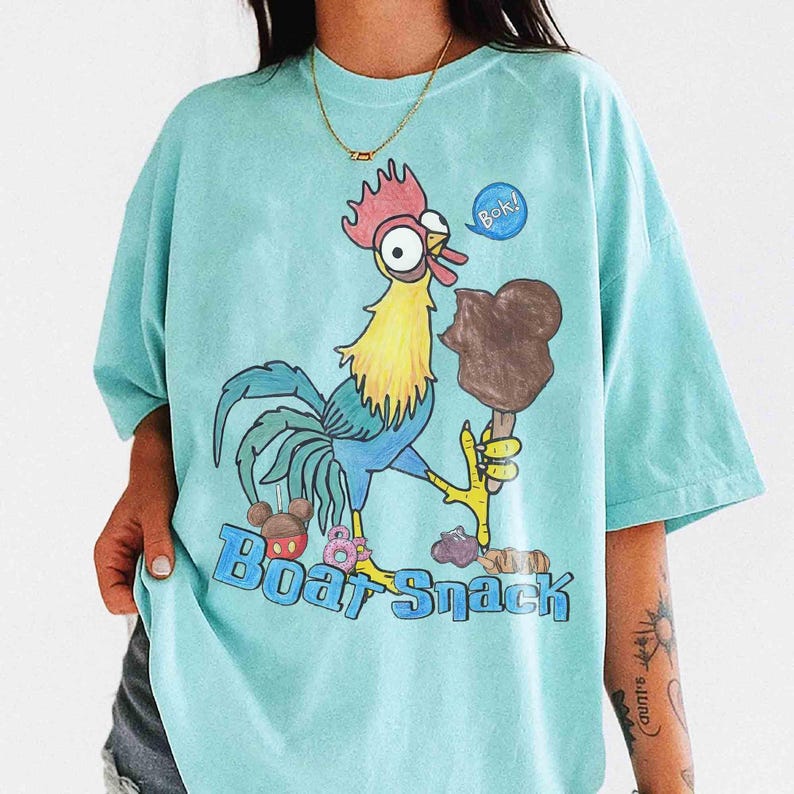 May include: A light blue t-shirt featuring a cartoon rooster holding a chocolate ice cream bar. The rooster has red, yellow, blue, and green details. Text on the shirt reads "Boa Snack" with a speech bubble saying "Bok!"