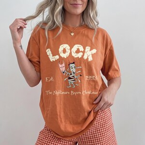 K&ouml;nnte beinhalten: Orangefarbenes Kurzarm-T-Shirt mit dem Wort "LOCK" in cremefarbener Schrift. Darunter eine Cartoon-Mumie, die eine Maske h&auml;lt, mit dem Text "Est. 1993" und "The Nightmare Before Christmas".