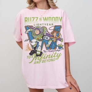 Może przedstawiać: R&oacute;żowa koszulka z nadrukiem przedstawiającym Buzza Astrala i Chudego z Toy Story. Projekt zawiera tekst "Buzz & Woody Lightyear" i "To Infinity and Beyond". Koszulka ma kr&oacute;tkie rękawy i okrągły dekolt.