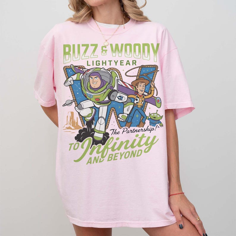 Może przedstawiać: R&oacute;żowa koszulka z nadrukiem przedstawiającym Buzza Astrala i Chudego z Toy Story. Projekt zawiera tekst "Buzz & Woody Lightyear" i "To Infinity and Beyond". Koszulka ma kr&oacute;tkie rękawy i okrągły dekolt.