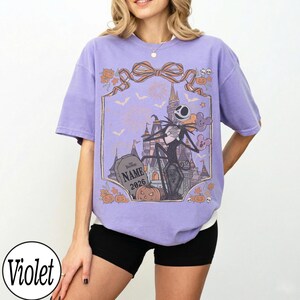 K&ouml;nnte beinhalten: Violettes T-Shirt mit einem Aufdruck von Jack Skellington vor einem Schloss, mit K&uuml;rbissen und Flederm&auml;usen. Das Design enth&auml;lt den Text "NAME 2026" auf einem Grabstein. Das Shirt hat eine lockere Passform.