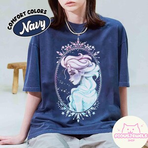 May include: Navy blue t-shirt with a watercolor illustration of a woman with long, flowing hair inside an oval frame. The shirt has the text "COMFORT COLORS Navy" on the upper left and the shop logo "PIOUSJEWELS SHOP" on the lower right.