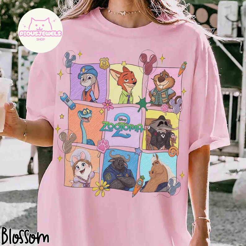 May include: Pink t-shirt featuring a colorful graphic of characters from the animated movie Zootopia. The design includes Judy Hopps, Nick Wilde, and other characters in a grid pattern. The text "Zootopia 2" is also visible.