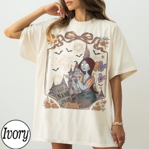 K&ouml;nnte beinhalten: Elfenbeinfarbenes T-Shirt mit einem Halloween-Motiv. Das Design zeigt eine Cartoonfigur, ein Schloss, Feuerwerk, Flederm&auml;use und K&uuml;rbisse. Die Grafik wird von einem dekorativen Rahmen mit einer Schleife oben umrahmt. Der Text "Happy Halloween NAME 2026" ist ebenfalls sichtbar.