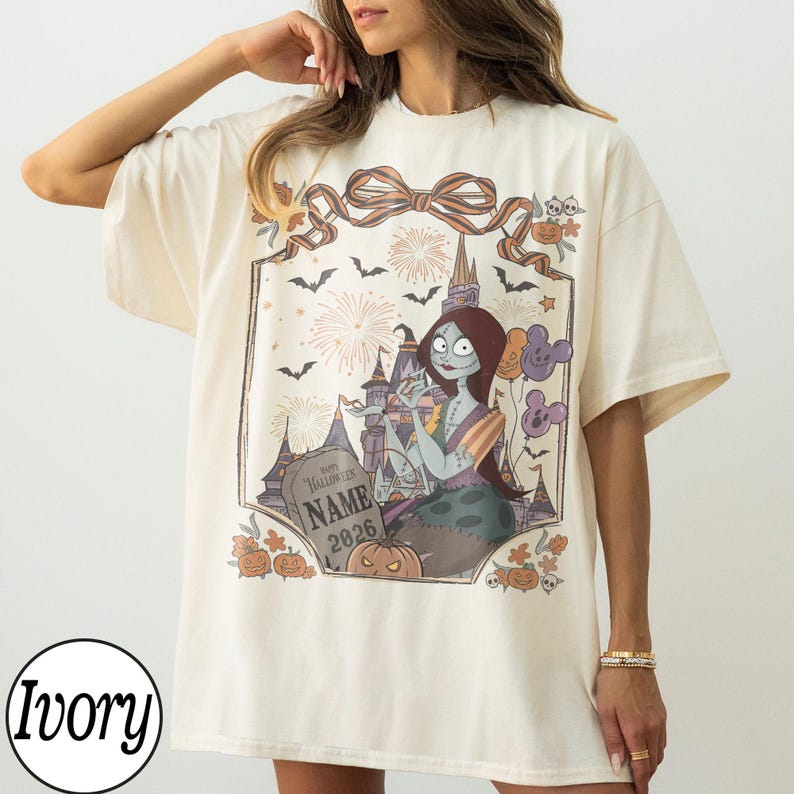 K&ouml;nnte beinhalten: Elfenbeinfarbenes T-Shirt mit einem Halloween-Motiv. Das Design zeigt eine Cartoonfigur, ein Schloss, Feuerwerk, Flederm&auml;use und K&uuml;rbisse. Die Grafik wird von einem dekorativen Rahmen mit einer Schleife oben umrahmt. Der Text "Happy Halloween NAME 2026" ist ebenfalls sichtbar.