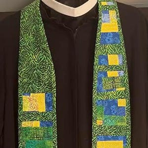 May include: A green and blue liturgical stole with a repeating pattern of yellow, blue, and green squares. The stole is made of fabric and is designed to be worn over the shoulders. The background is a black garment.