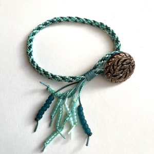 May include: A green and white braided bracelet with a green and white tassel and a gold button with a leaf design.