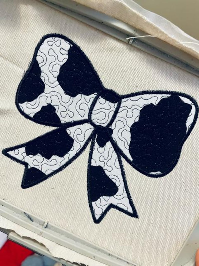 May include: A black and white cow print bow tie embroidery design on a white fabric.