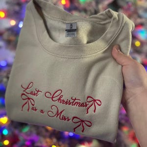 May include: A beige sweatshirt with the text "Last Christmas as a Miss" embroidered in red with a bow design.
