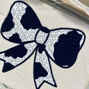 May include: A black and white cow print bow tie embroidery design on a white fabric.