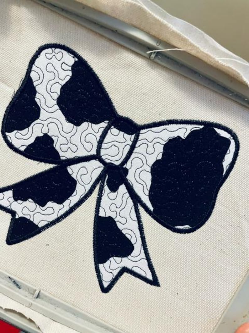 May include: A black and white cow print bow tie embroidery design on a white fabric.