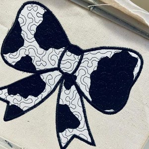May include: A black and white cow print bow tie embroidery design on a white fabric.