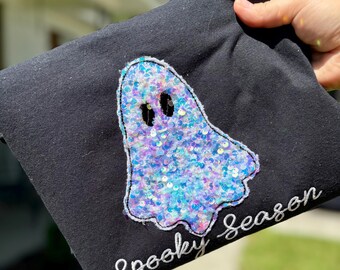May include: Black sweatshirt featuring a sequined ghost design in shades of blue, purple, and pink. The ghost has two black eye cutouts. The words "Spooky Season" are embroidered in white script below the ghost.