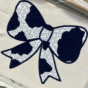 May include: A black and white cow print bow tie embroidery design on a white fabric.