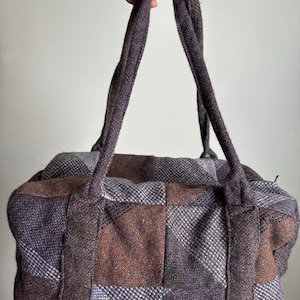 以下が含まれることがあります： 茶色、グレー、紫の色合いのパッチワークダッフルバッグ。バッグには2つの頑丈なハンドルと長方形の形状があります。生地は、キルティングデザインの、質感のあるツイードまたは同様の素材のようです。