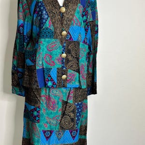 Op de afbeelding: Een tweedelige outfit met een lang mouwjack en een rok. De stof is een patchwork van blauw, groen, zwart en roze met gouden accenten. Het jack heeft een V-hals en gouden knopen.