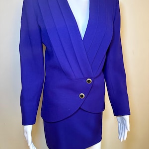 Op de afbeelding: Een levendig paars tweedelig pak. De blazer heeft een V-hals met geplooide details en goudkleurige knopen. De bijpassende rok is een klassieke, knielange stijl. Het pak wordt op een mannequin getoond.