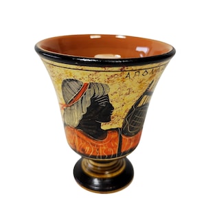 Può includere: Un vaso di ceramica con un design nero e arancione. Il vaso presenta il profilo di una persona con capelli neri e una fascia rossa. Il vaso ha la scritta "ΑΠΟΛ". L'interno del vaso è arancione.