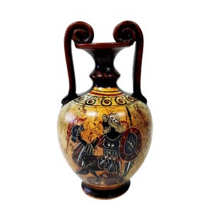 Puede incluir: Un jarrón decorativo de estilo antiguo con una ilustración detallada de figuras griegas antiguas. El jarrón presenta una base y asas de color marrón oscuro, con un cuerpo amarillo y marrón. La obra de arte representa a un guerrero con lanza y escudo.