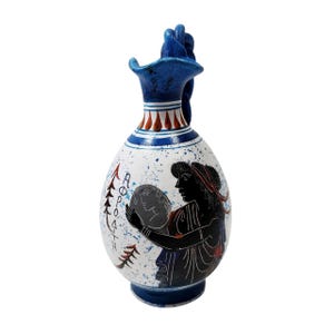 May include: A ceramic vase with a blue handle and rim, featuring a white body with blue speckles. The vase has a black illustration of a woman holding a round object, with Greek text. Red and brown accents are present.