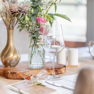 May include: A table setting with a wooden place card that says "Michael" on a wine glass, a gold vase with dried flowers, and a clear glass vase with pink flowers.