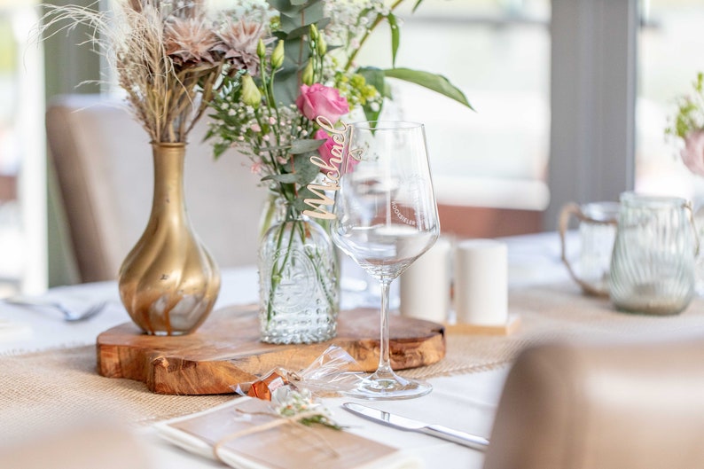 May include: A table setting with a wooden place card that says "Michael" on a wine glass, a gold vase with dried flowers, and a clear glass vase with pink flowers.
