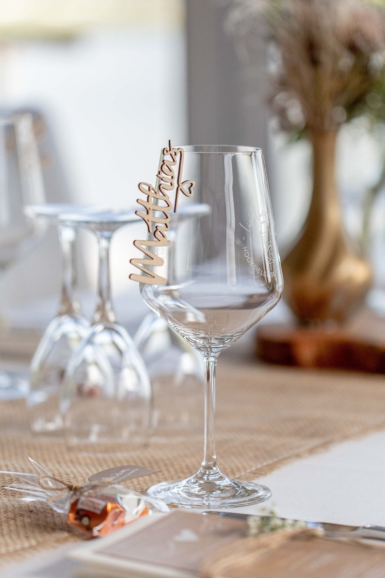 May include: A clear wine glass with a wooden place card that says "Matthias" on it. The glass is sitting on a table with a brown burlap runner.