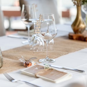 May include: A rustic wedding table setting with a burlap runner, a clear glass candle holder, a small gift wrapped in burlap, a white napkin, a menu, a silver fork, a silver knife, and a wine glass with a wooden name card that says "Matthias".