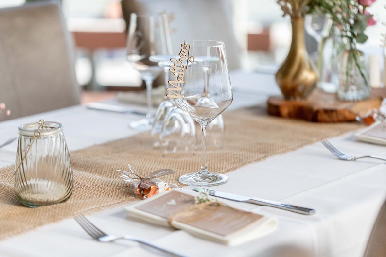May include: A rustic wedding table setting with a burlap runner, a clear glass candle holder, a small gift wrapped in burlap, a white napkin, a menu, a silver fork, a silver knife, and a wine glass with a wooden name card that says "Matthias".