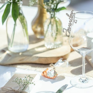 May include: A clear glass wine glass with a wooden name card that says "Jennifer" attached to the rim. The glass is on a white tablecloth with a small brown gift box and a sprig of white flowers.
