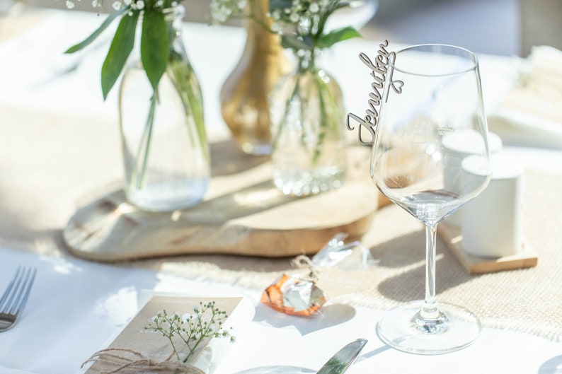 May include: A clear glass wine glass with a wooden name card that says "Jennifer" attached to the rim. The glass is on a white tablecloth with a small brown gift box and a sprig of white flowers.