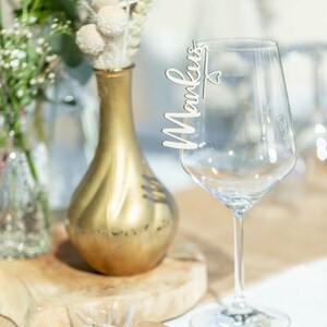 May include: A wooden place card with the name "Markus" written on it is attached to a clear wine glass. The glass is sitting on a table with a brown burlap runner and a small gift bag with a heart-shaped tag that says "Sch&ouml;n dass du da bist!"