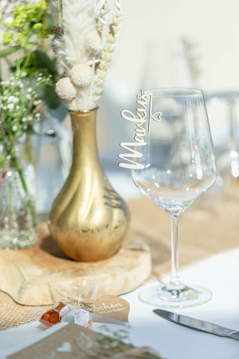 May include: A wooden place card with the name "Markus" written on it is attached to a clear wine glass. The glass is sitting on a table with a brown burlap runner and a small gift bag with a heart-shaped tag that says "Sch&ouml;n dass du da bist!"