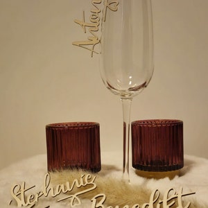 May include: Wooden place cards with names written in cursive, including "Stephanie", "Vanessa", "Benedikt", and "Julian". A champagne flute with a wooden place card that says "Antonio" is in the background. The place cards are on a white fluffy surface.
