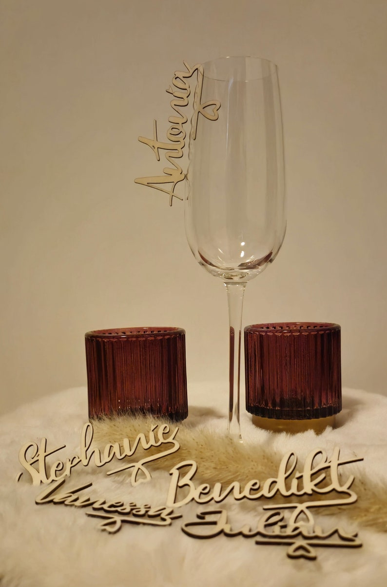 May include: Wooden place cards with names written in cursive, including "Stephanie", "Vanessa", "Benedikt", and "Julian". A champagne flute with a wooden place card that says "Antonio" is in the background. The place cards are on a white fluffy surface.