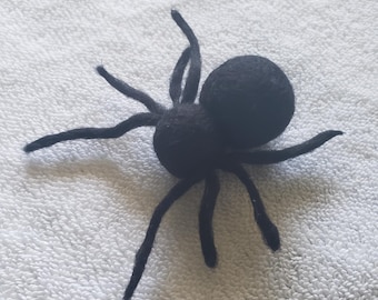 Puede incluir: Una araña de fieltro negro con ocho patas y un cuerpo redondo. La araña está sentada sobre una superficie blanca.