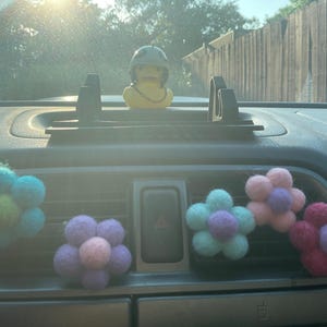 Peut inclure: Cinq désodorisants de voiture en forme de fleurs en feutre dans l'aérateur d'une voiture. Les fleurs sont bleues, roses, violettes, vertes et rouges.