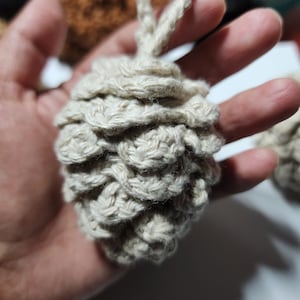 Puede incluir: Adorno de piña tejido a ganchillo en color beige natural. El adorno hecho a mano presenta un diseño texturizado en forma de escamas y un lazo para colgar. Ideal para decoración festiva o rústica.