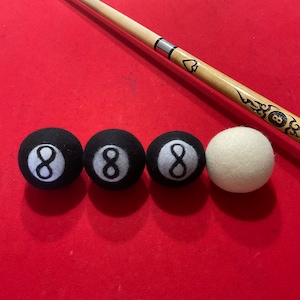 Peut inclure: Quatre boules de billard noires avec le numéro huit imprimé en blanc sur chaque boule, et une boule de billard blanche, le tout sur une surface de table de billard en feutre rouge. Une queue de billard en bois avec un motif noir et blanc est posée sur la table.