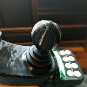 Könnte beinhalten: Nahaufnahme eines schwarzen Joysticks mit kugelförmigem Griff. Der Joystick ist Teil eines Bedienfelds mit weißen Knöpfen und einem grün beleuchteten Streifen. Der Griff hat eine strukturierte, geriffelte Oberfläche.